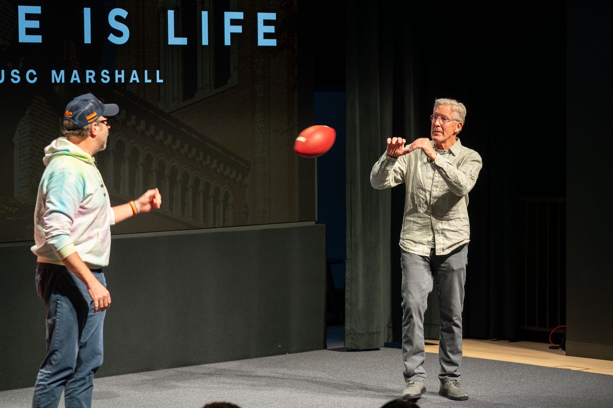 Jason Sudeikis and Pete Carroll standing on stage with the Game is Life USC Marshall banner behind them